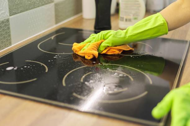 Person wearing green gloves cleaning a glass stove top with an orange cloth and soapy water.